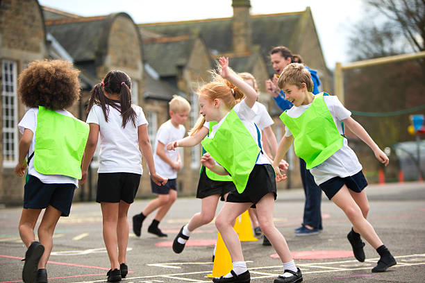 Children running around in the school playground during a physical education lesson. Some are wearing coloured vests and there are yellow cones on the floor that they are using for the task. The teacher is cheering them on and the childring are laughinas they run around.