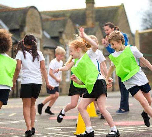 Children running around in the school playground during a physical education lesson. Some are wearing coloured vests and there are yellow cones on the floor that they are using for the task. The teacher is cheering them on and the childring are laughinas they run around.